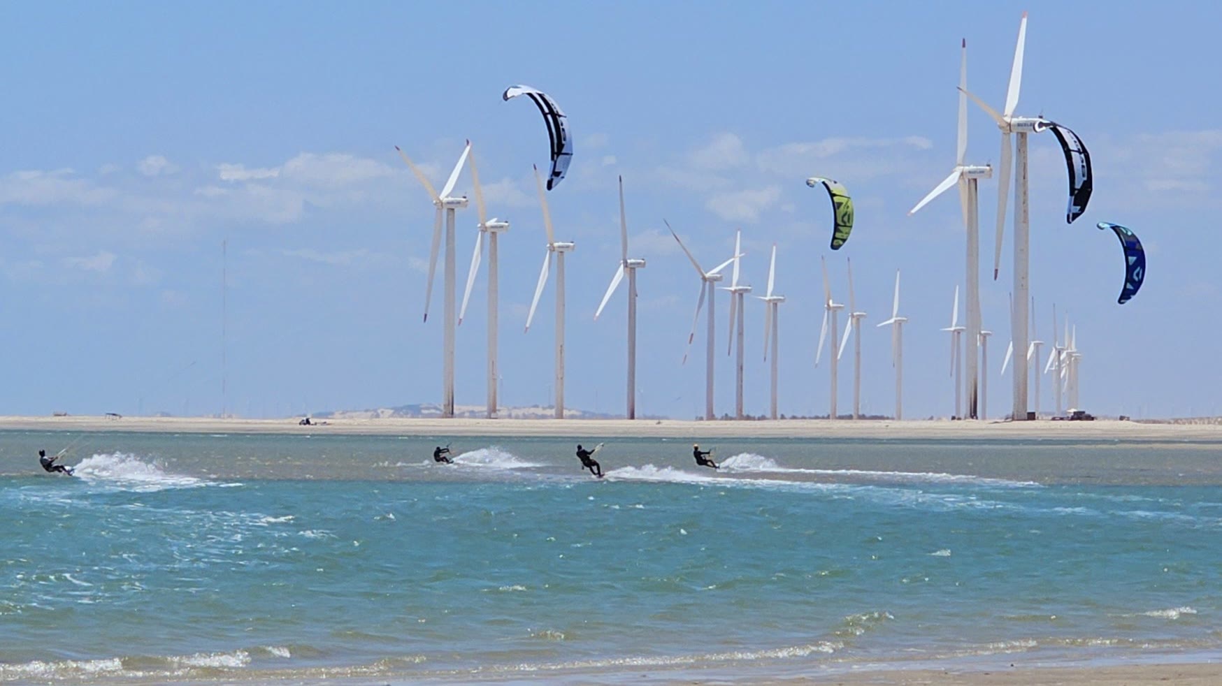 Kitesurfers riding in front of a wind farm, kites high in the sky